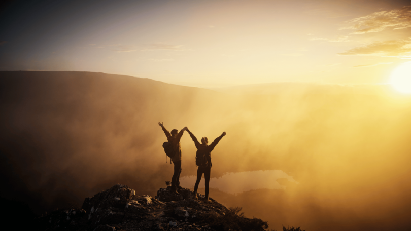 Imagem de uma montanha e o pôr do Sol. Em destaque um casal de mãos dadas e braços levantados, agradecendo a oportunidade de estarem vivos e vivendo a vida.