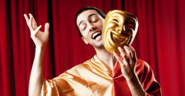 Imagem de um ator masculino em cima do palco de um teatro. Ele está de olhos fechados, sorrindo e segurando em uma das mãos uma máscara dourada.