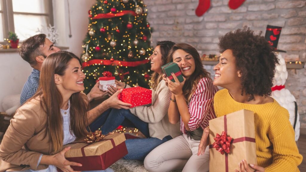 Imagem de vários amigos e amigas reunidos em uma sala decorada com vários artigos natalinos, trocando seus presentes.
