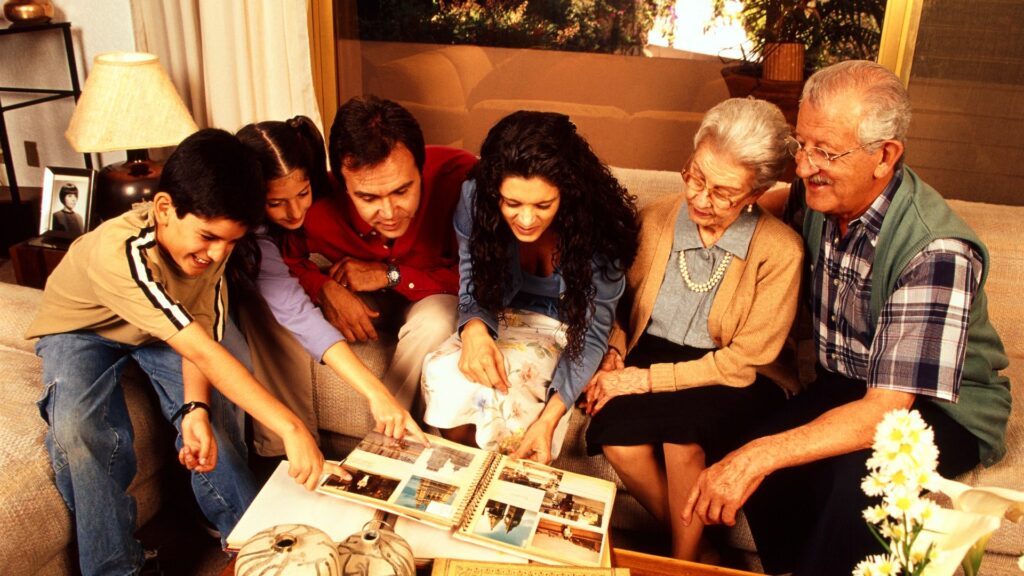 Imagem de uma família, avós, pais e filhos, reunidos em uma sala, vendo um álbum de fotografias.
