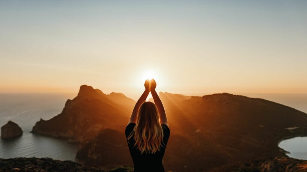 Imagem de uma montanha ao fundo com o pôr do Sol e em destaque uma mulher segurando o Sol, simbolizando a renovação espiritual.