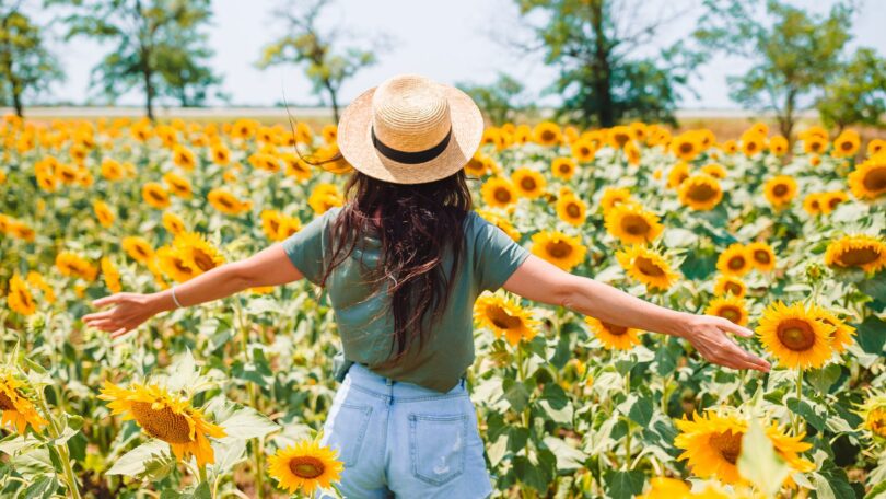 Imagem de uma jovem usando um chapéu de palha dentro de um campo de girassóis curtindo a natureza.