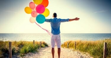 Imagem de fundo de um lindo mar azul e em destaque um homem usando uma bermuda branca e um camisa jeans. Ele está contemplando a beleza do mar e a felicidade da vida. Ele ainda está segurando em uma das mãos, vários balões coloridos.