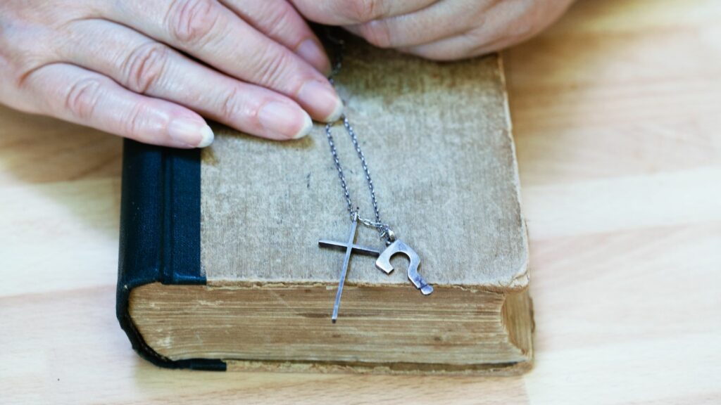 Imagem de um bíblia antiga sobre uma mesa de madeira. Mãos de uma mulher seguram uma corrente com um crucifixo e um pingente com ponto de interrogação, simbolizando a existência ou não de Deus.
