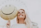 Imagem de uma mulher loura, de cabelos longos, sorrindo. Ela está usando uma blusa rendada na cor branca segurando um balão com uma carinha de sorriso, trazendo o conceito de feicidade.