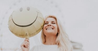 Imagem de uma mulher loura, de cabelos longos, sorrindo. Ela está usando uma blusa rendada na cor branca segurando um balão com uma carinha de sorriso, trazendo o conceito de feicidade.