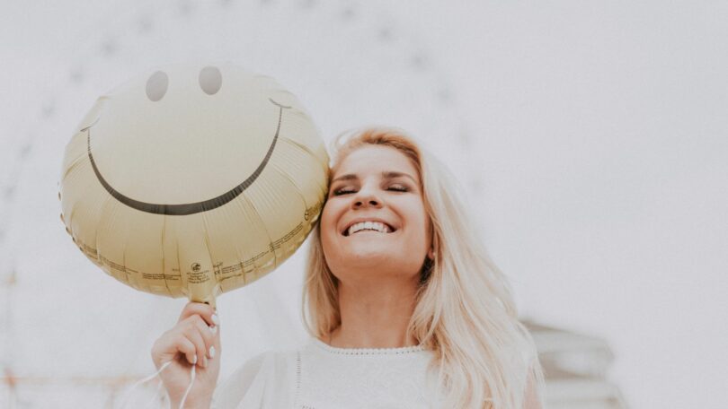 Imagem de uma mulher loura, de cabelos longos, sorrindo. Ela está usando uma blusa rendada na cor branca segurando um balão com uma carinha de sorriso, trazendo o conceito de feicidade.