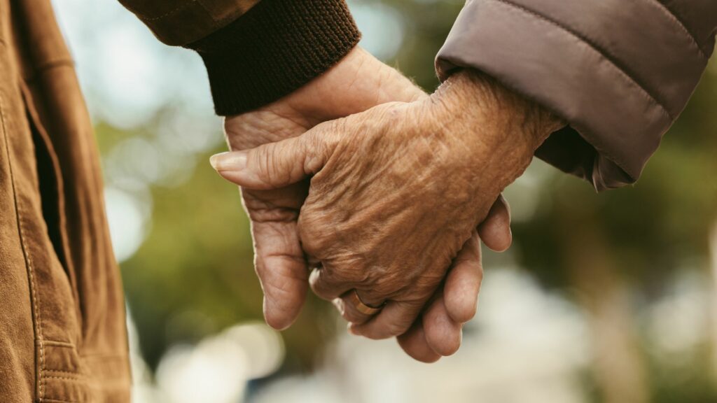 Imagem de um casal de idosos de mãos dadas caminhando em um jardim.
