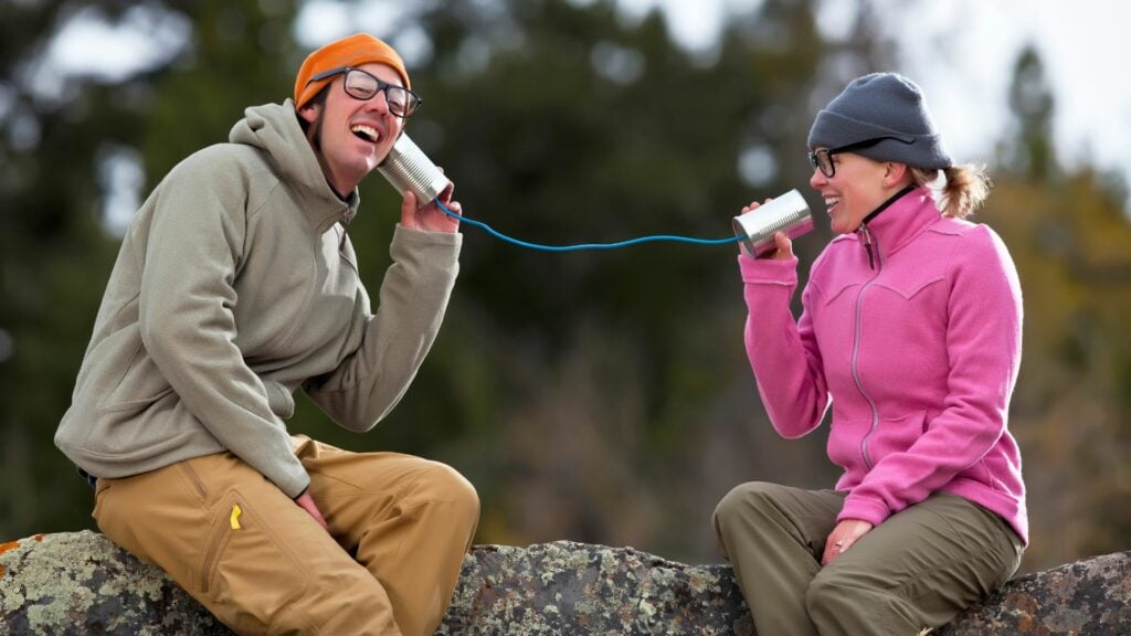 Imagem de um casal sentado em uma pedra, ambos estão usando roupas de inverno e se comunicando por meio de duas latas e um fio, simbolizando o conceito de comunicadores.
