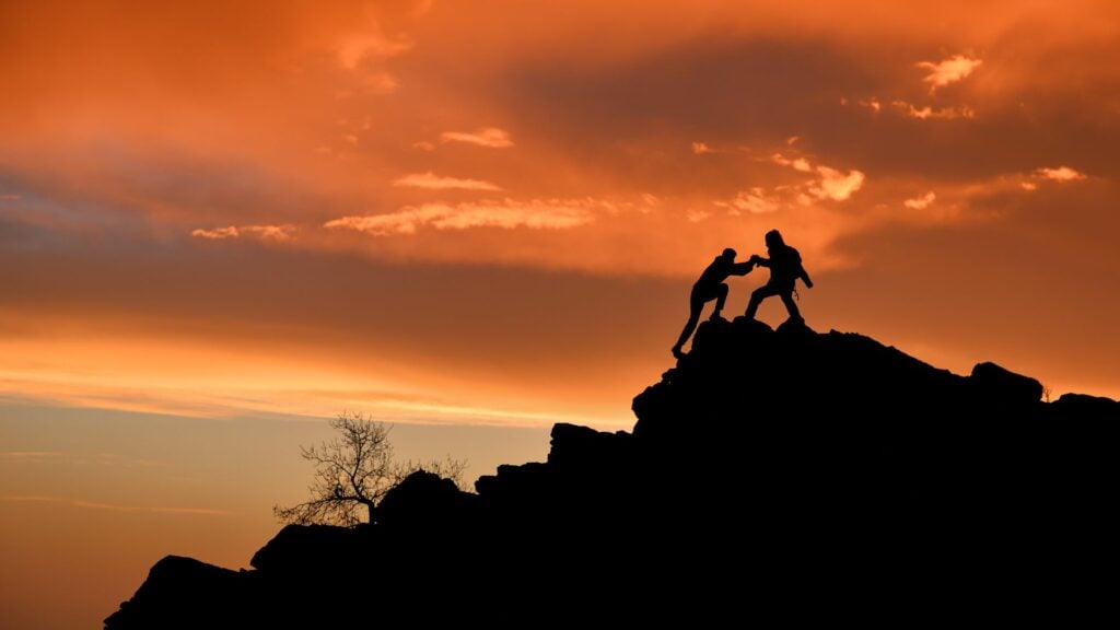 Imagem de um lindo por do sol e em destaque duas pessoas escalando uma montanha, simbolizando o conceito de superação de desafios por meio da espiritualidade.
