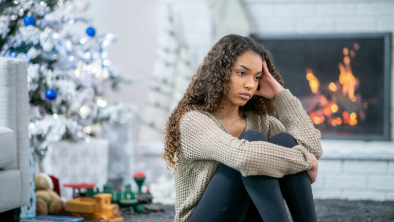 Imagem de uma sala decorada para o Natal e festas de final de ano. Em destaque, a foto de uma menina de cabelos longos e cacheados, sentada no chão com o semblante triste.