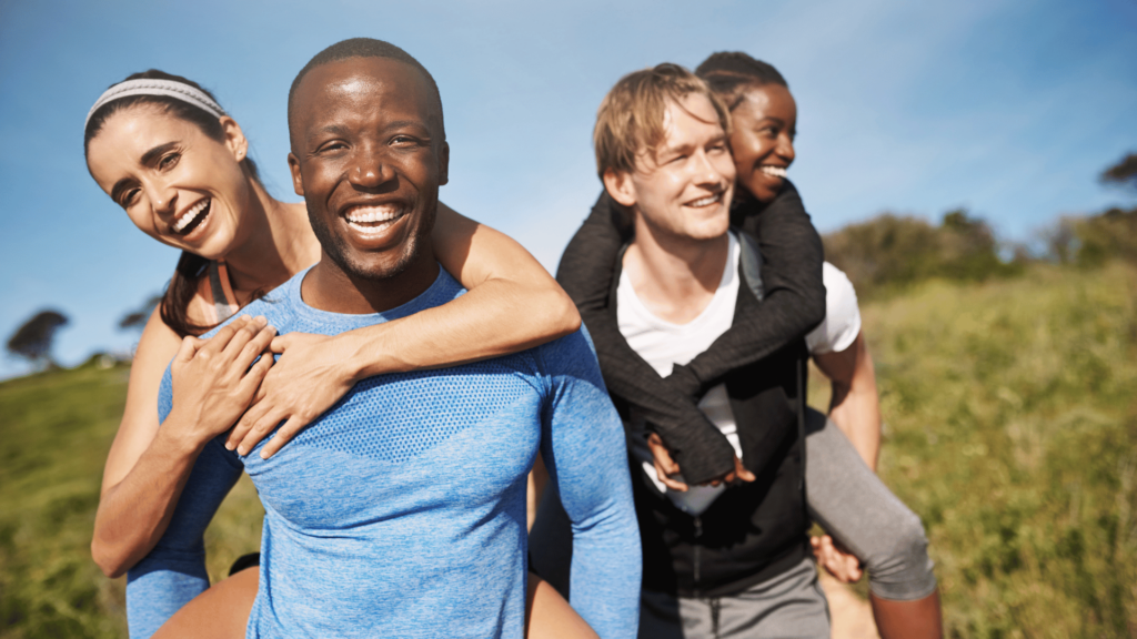 Imagem de dois casais, 2 homens e duas mulheres, andando por uma estrada de terra. Os homens carregam as mulheres nas costas, em tom de alegria e bricandeira. Todos estão sorrindo e felizes.
