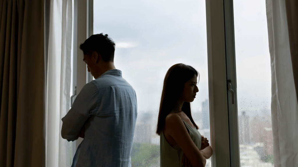 Um casal está de costas um para o outro, provavelmente estão bravos após uma discussão. Ao fundo, há uma janela aberta e uma cidade desfocada.