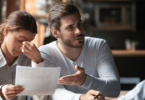 Um casal está em uma mesa. A mulher está com uma mão no rosto e tem a expressão de cansaço. Ao seu lado, o homem está reclamando com a pessoa na frente deles.