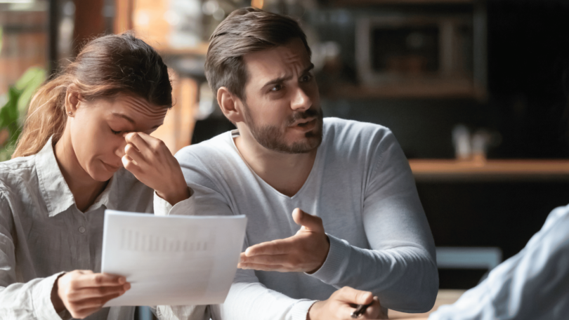 Um casal está em uma mesa. A mulher está com uma mão no rosto e tem a expressão de cansaço. Ao seu lado, o homem está reclamando com a pessoa na frente deles.