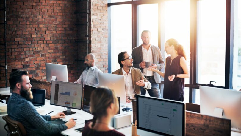 Imagem de um ambiente corporativo, uma sala, com vários computadores e pessoas sentadas e em pé, conversando.