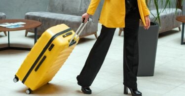 Imagem de uma mulher usando uma roupa preta e um casaco amarelo, puxando uma mala de rodinhas na cor amarela.