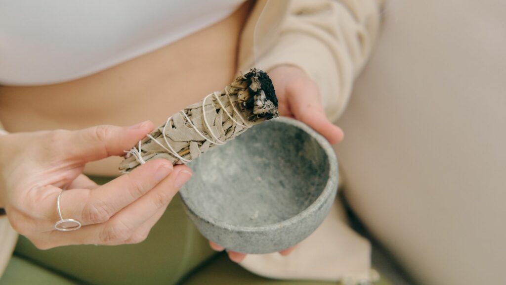 Imagem das mãos de uma mulher. Em uma delas ela segura um bowl de cerâmica e na outra um incenso, trazendo o conceito de práticas holísticas.
