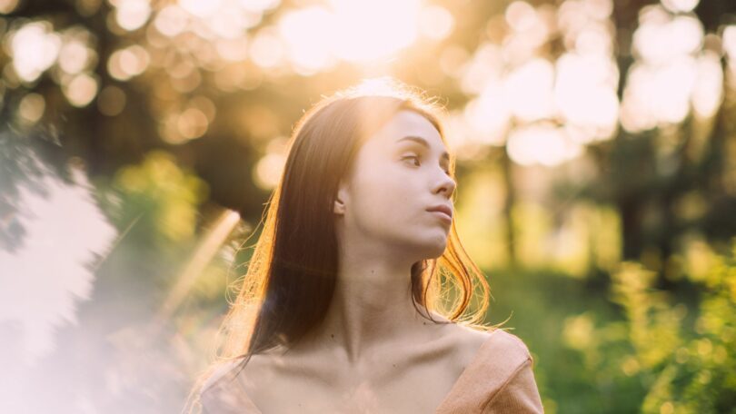 Imagem ao ar livre de uma jovem no fundo da natureza. Ela está reflexiva e calma. A foto traz o conceito de Medicina Holística e Integrativa, Terapias Mente-Corpo e Técnicas de Relaxamento.