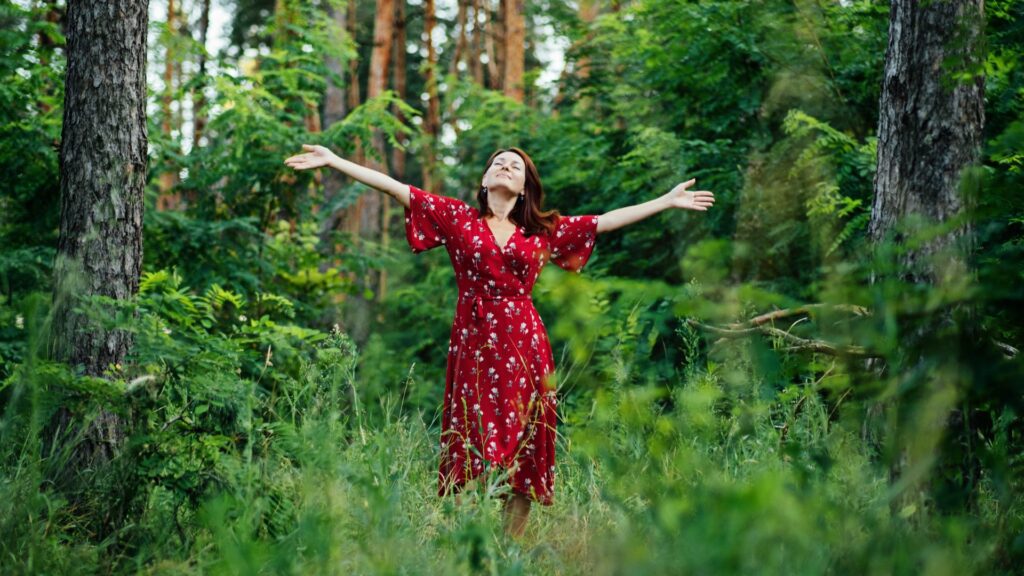 Imagem de uma floresta e em destaque uma mulher usando um vestido vermelho florido. Ela está de braços abertos, trazendo o conceito de conexão com a natureza.
