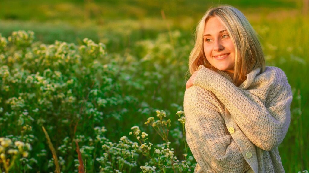 Imagem de uma moça jovem usando uma blusa de lã bege. Ela está sorridente em um campo de flores, trazendo o conceito de transformação interna, de feridas cicatrizadas.
