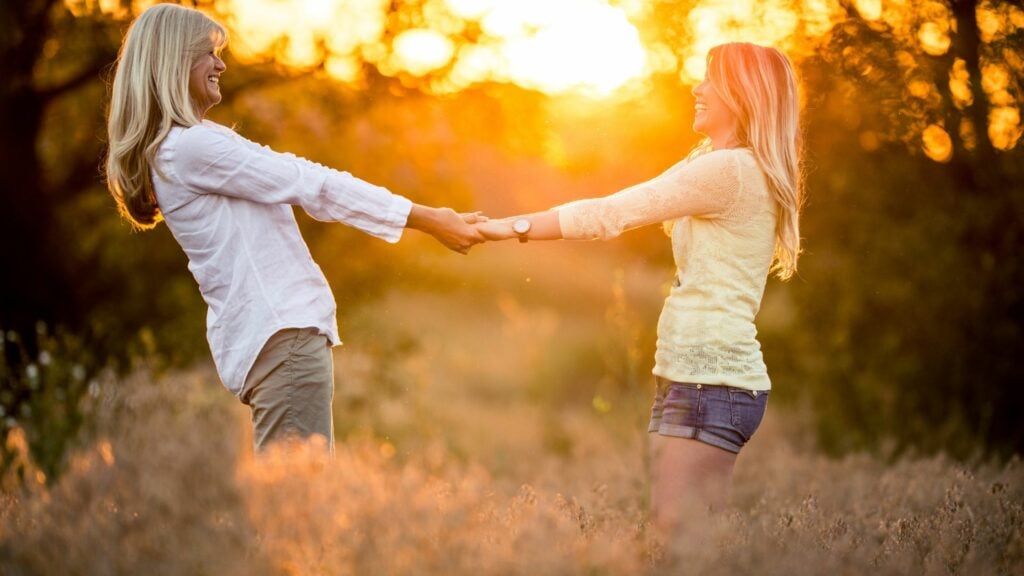 Imagem de duas mulheres de mãos dadas em um campo. Ao fundo um lindo pôr do Sol. Elas estão felizes e trazem o conceito que uma é importante para a outra.
