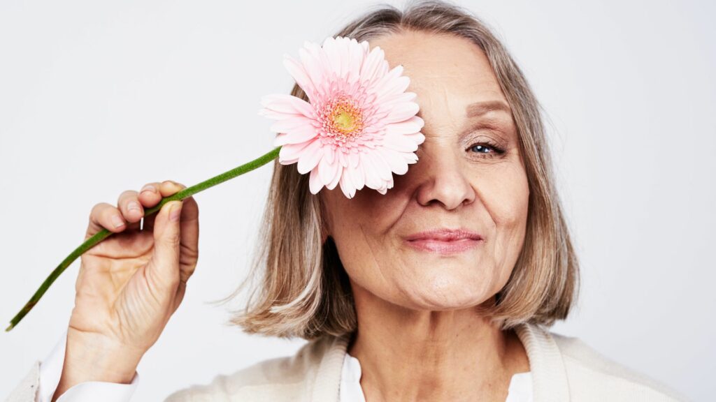 Imagem de uma mulher idosa, segurando em uma das mãos uma flor rosa clara. A imagem, leve, traz o conceito de cuidado e polimento da alma, após a terceira idade, o preparo para o retorno ao plano espiritual.
