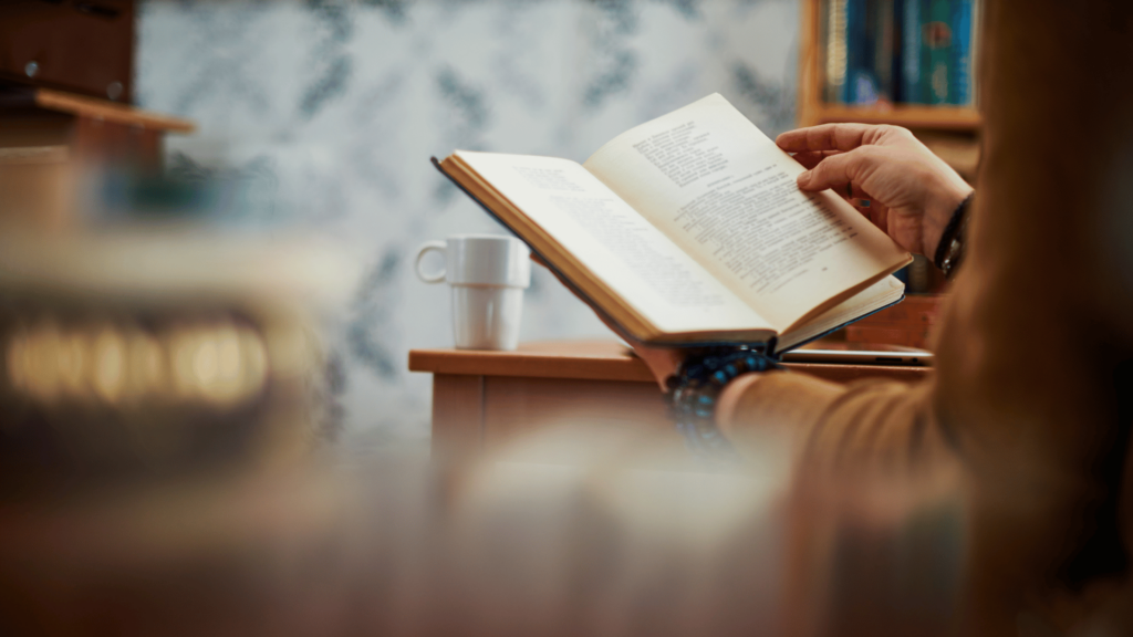 As mãos de uma pessoa estão no foco na imagem e elas seguram um livro aberto. Há uma xícara em cima de uma mesa.