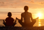 Mãe e filho estão sentados de costas para a imagem e meditam de frente para o mar e para o pôr do sol.