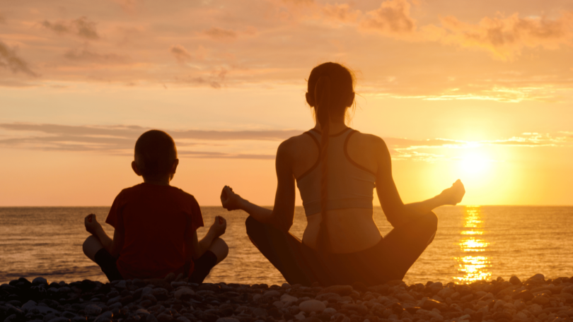 Mãe e filho estão sentados de costas para a imagem e meditam de frente para o mar e para o pôr do sol.