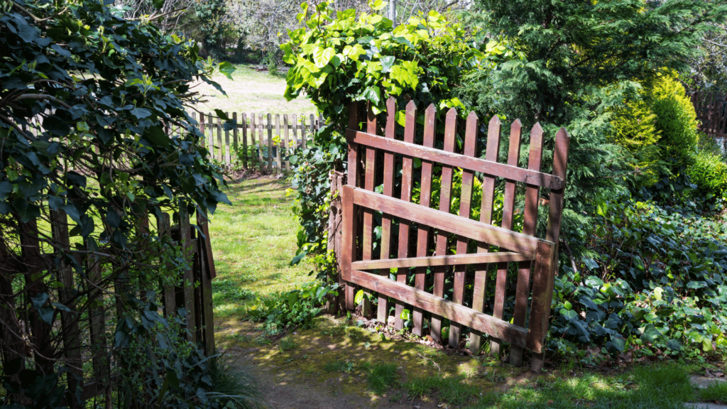 Um portão de madeira aberto que leva para um jardim ou um ambiente de natureza. Há muitas plantas e folhas ao redor.