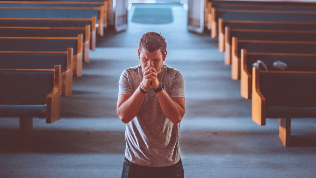 Um homem está na frente de um altar em uma igreja. Ele junta as duas mãos e reza.