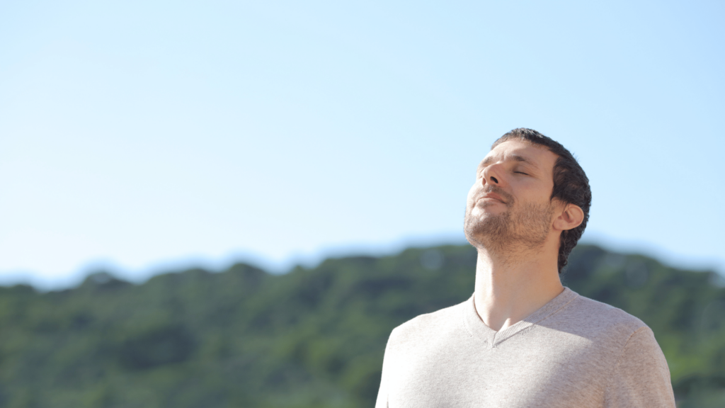 Um homem que está ao ar livre levanta a cabeça e respira o ar fresco. Atrás dele, há montanhas e o céu azul.