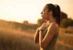 Uma mulher jovem está ao ar livre. Ela respira o ar fresco, está com os olhos fechados, junta as duas mãos e as coloca sobre o peito, como se estivesse meditando na natureza.