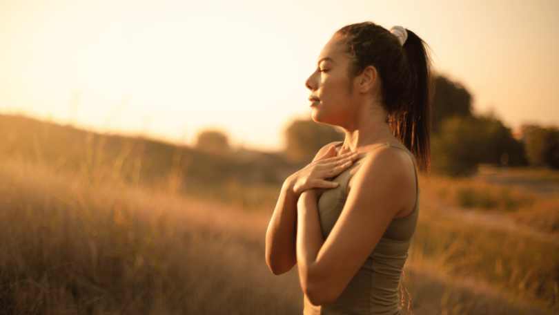 Uma mulher jovem está ao ar livre. Ela respira o ar fresco, está com os olhos fechados, junta as duas mãos e as coloca sobre o peito, como se estivesse meditando na natureza.