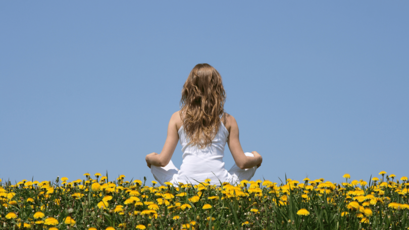 Uma mulher está sentada de costas para a imagem ao ar livre. Ela faz a posição da meditação e observa um céu azul claro. Ela está sentada em um campo cheio de flores amarelas.