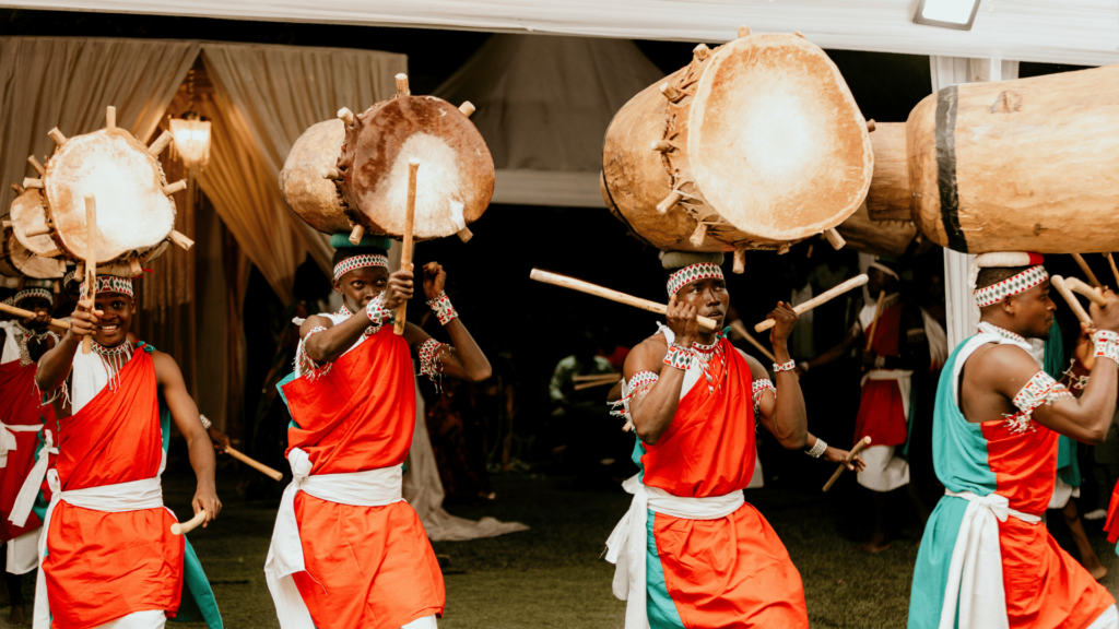 Quatro homens africanos estão no foco da imagem. Eles fazem uma apresentação ou dança típica da tradição do seu povo e carregam tambores sobre suas cabeças.