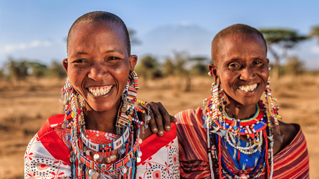 Duas mulheres da tribo Masai do Quênia estão no foco da imagem. Elas estão sorrindo e usam roupas e colares coloridos.