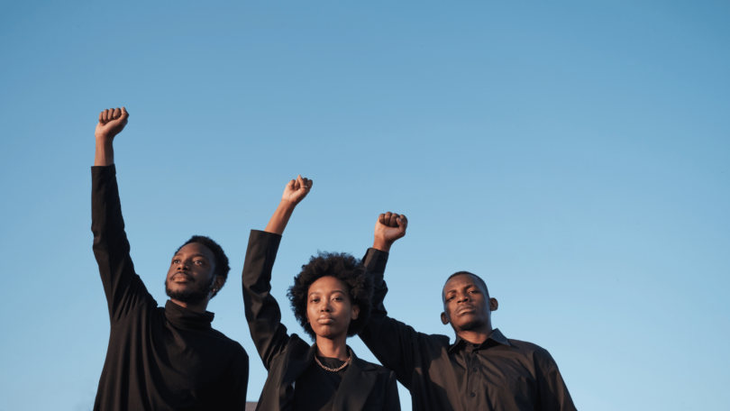 Três pessoas negras, uma mulher e dois homens, estão no foco da imagem. Eles levantam os seus punhos das mãos direitas no ar. Eles usam roupas pretas. Ao fundo, há um céu azul claro.