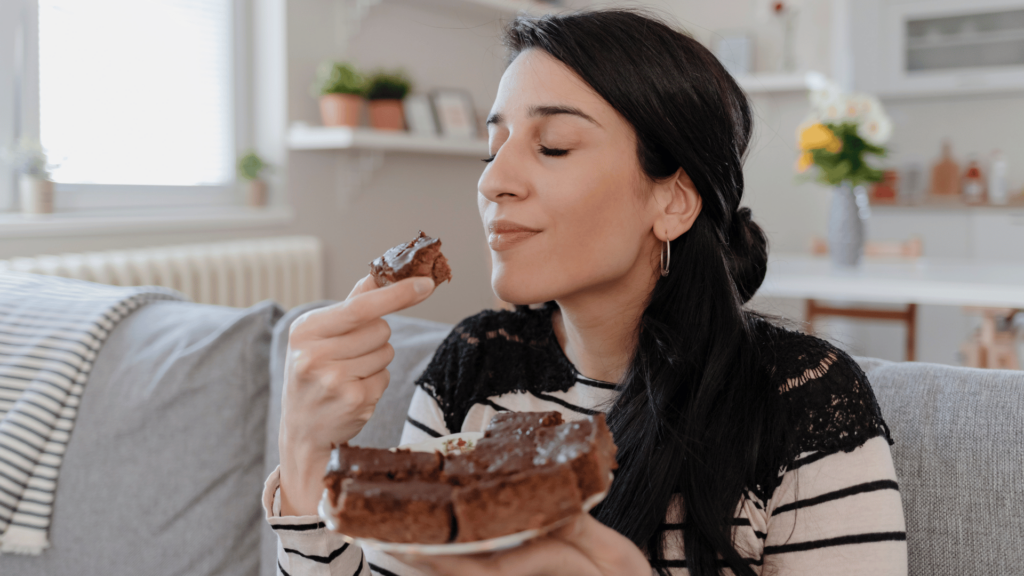 Uma mulher comendo um bolo de chocolate está no foco da imagem. Ela fecha os olhos e leva um pedaço do bolo à boca. Em outra mão, ela segura um prato com mais pedaços de bolo.