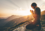 Um homem ajoelhado com as duas mãos unidas em sinal de oração está no foco da imagem. Ele está com os olhos fechados e a cabeça inclinada para baixo. Ele está em um ambiente de natureza, em cima de uma montanha. Ao fundo, há montanhas e o pôr do sol.