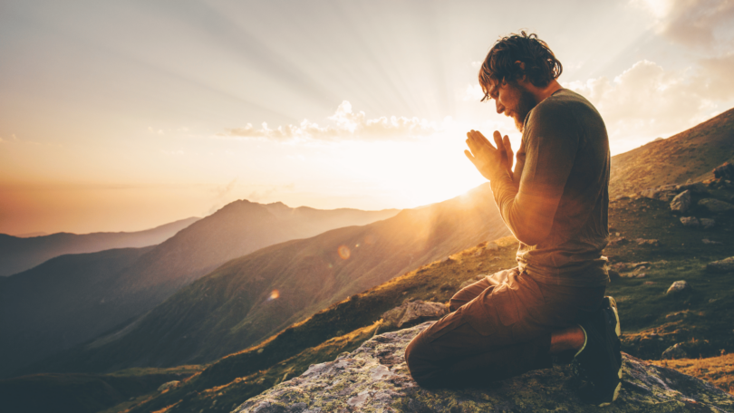 Um homem ajoelhado com as duas mãos unidas em sinal de oração está no foco da imagem. Ele está com os olhos fechados e a cabeça inclinada para baixo. Ele está em um ambiente de natureza, em cima de uma montanha. Ao fundo, há montanhas e o pôr do sol.