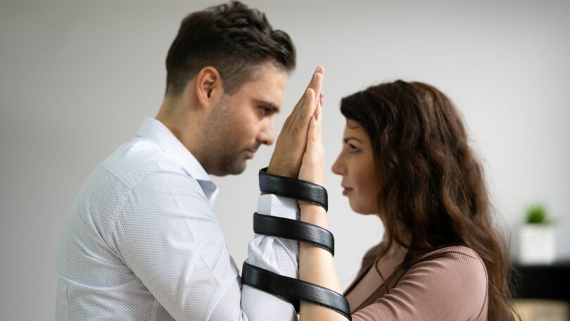 Imagem de um casal olhando um para o outro de mãos levantadas, tendo os braços entrelados por uma fita preta, trazendo o conceito de uma relação tóxica.