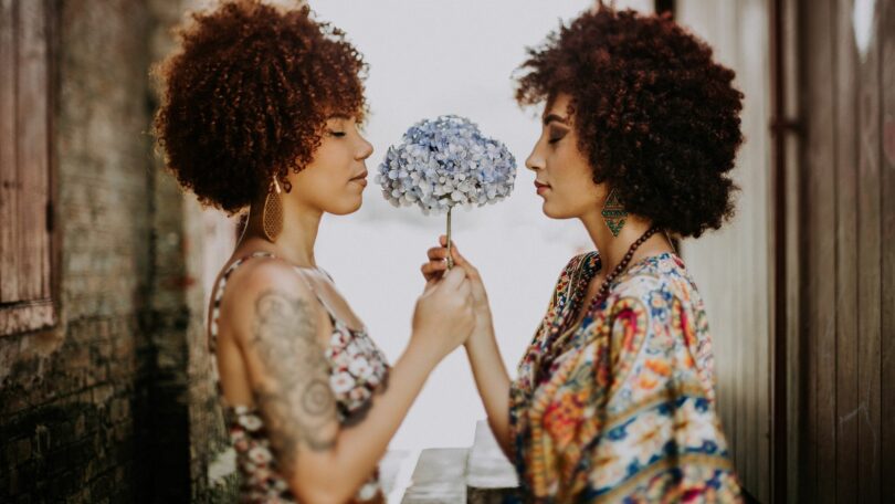 Imagem de duas mulheres frente a frente segurando uma flor de pétalas brancas e azuis, trazendo o conceito de compartilhar.