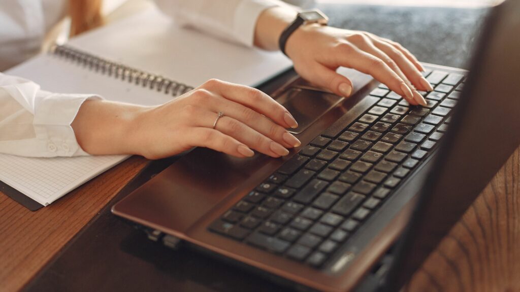 Imagem da mãos de uma mulher digitando em seu notebook. Ela está em seu ambiente de trabalho usando as novas tecnologias para ajudar nas tarefas do cotidiano.
