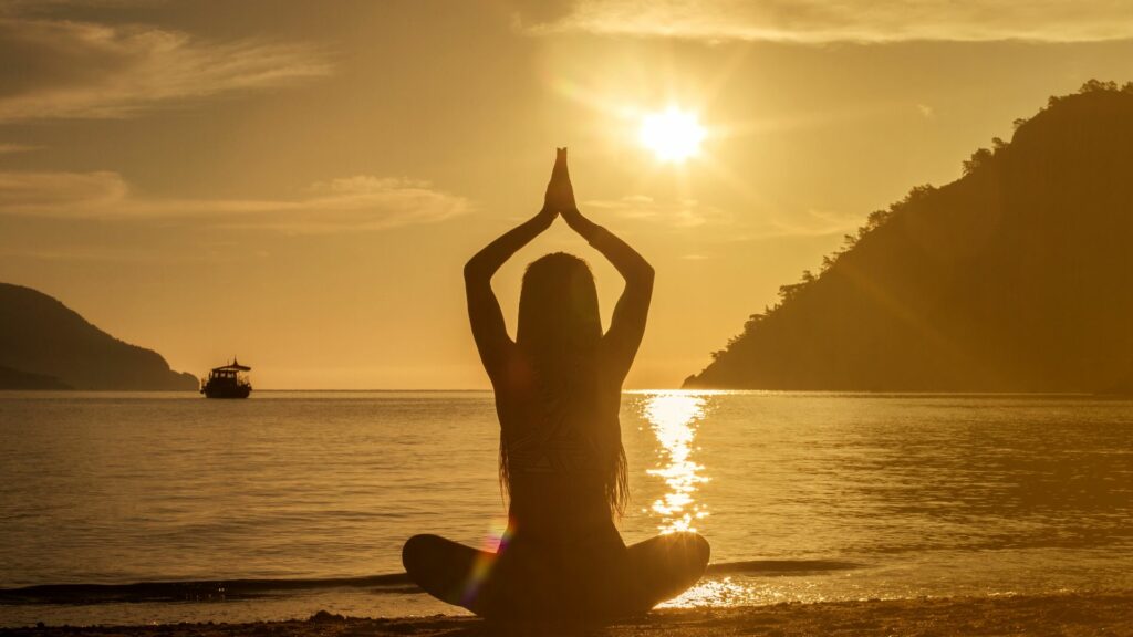 Imagem do pôr do sol em uma praia e em destaque uma mulher sentada com os braços erguidos fazendo uma meditação, trazendo o conceito de evolução espiritual.