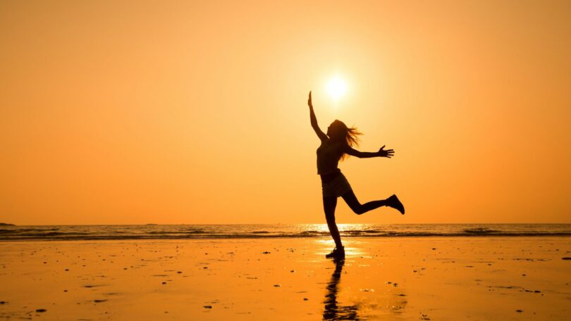 Imagem do pôr do sol em uma praia e em destaque a silhueta de uma moça feliz e radiante, trazendo o conceito de uma pessoa iluminada.