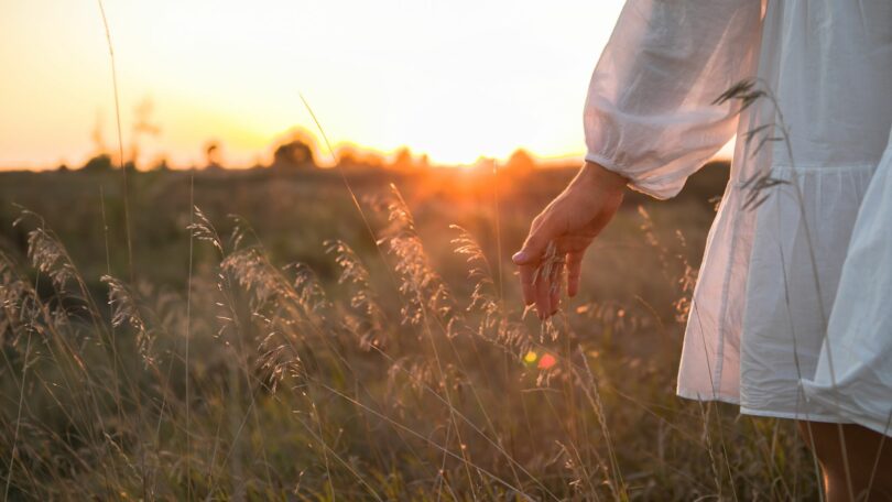 Imagem de um campo e em destaque uma mulher usando um vestido branco. Na foto, aparece apenas uma parte dela e sua mão esquerda.