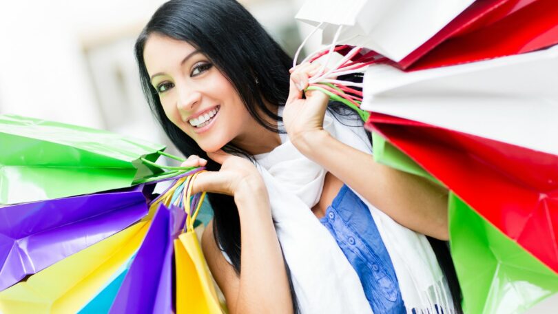 Imagem de uma jovem mulher, com cabelos longos e preto, segurando várias sacolas coloridas, trazendo o conceito de compras por compulsão.