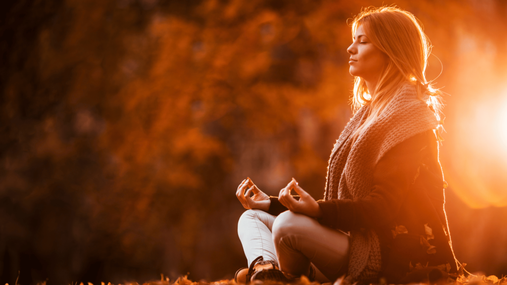 Uma mulher sentada e que pratica a meditação está no foco da imagem. Ela está com os olhos fechados e as pernas cruzadas. No fundo, há um ambiente de natureza e luz do pôr do sol.